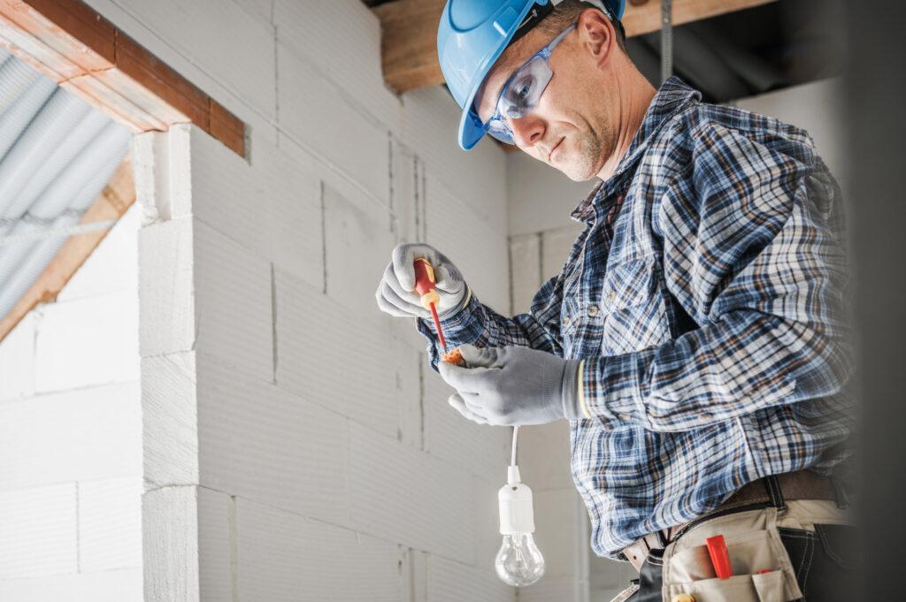 An adult electrician wearing a hard hat, protective eyewear, gloves, and a plaid shirt is seen working on connecting wires. He holds a screwdriver in one hand and the lightbulb component in the other. He is wearing a tool belt. The location is an indoor construction site with unfinished walls made of white bricks. This image evokes a mood of focused and skilled labor and could be used for projects related to construction, home improvement, skilled trades, or the electrical industry.